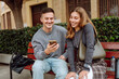 © maxbelchenko - A young woman and a man are sitting on a bench with a phone in a city park. Friends are chatting outdoors, looking at their phones at sunset. Concepts of friendship, communication, and technology.