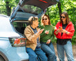 © olezzo - Happy senior mother and two adult daughters sitting in open car trunk and looking at smartphone