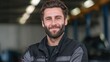 © standret - Caucasian male auto mechanic with beard smiles confidently while standing in a garage, tools and vehicles visible in the background, wearing black work uniform