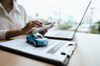 © Natthawadee - A woman reviews and signs a car insurance policy at her desk with a calculator, laptop, and miniature cars, representing auto coverage planning and financial protection.