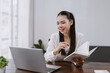 © amnaj - Young Asian woman holding a notebook and pen, engaging in e learning or remote work on a laptop in a home office