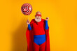 © deagreez - Senior superhero in red cape blue suit holds giant lollipop with smile against bright yellow background