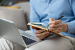 © Liubomir - Man holding an open notebook and writing notes with a pen while sitting with a laptop on his lap, focusing on work, planning, and remote learning concepts