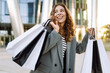 © maxbelchenko - A cheerful woman in stylish clothes with shopping bags in front of a modern building. A stylish woman enjoys a sunny day after shopping. Concept of consumerism, shopping.
