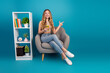 © deagreez - Young woman in a chic casual setting sits on a stylish chair with a smartphone in a bright blue studio