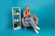 © deagreez - Young woman sits in a comfy chair using smartphone with bright blue studio background and decorative bookshelf