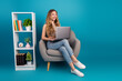 © deagreez - Young chic woman sits in a cozy chair with a laptop beside a bookshelf against a vibrant blue background for lifestyle fashion and study themed image