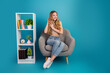 © deagreez - Young chic woman relaxing in a stylish armchair beside shelf with books plants in a bright blue studio