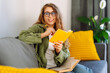 © maxbelchenko - Young woman sits on sofa with yellow cushions and writes in a yellow notebook. Smiling woman relaxes in cozy room, taking notes, enjoying some alone time. Concepts of comfort, education, relaxation.