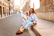 © maxbelchenko - Portrait of a happy woman with curly hair, holding phone, strolling along sunny city streets. Beautiful tourist in sunglasses, holding phone enjoys sunny day and architecture. Tourism concept, urban.