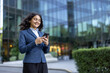 © Liubomir - Young businesswoman standing outdoors in front of a modern glass building, actively using her smartphone and smiling, embodying communication and professional connectivity