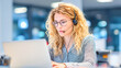 © Kowit - A young woman with curly blonde hair and glasses works attentively on a laptop, wearing a headset in a modern, softly lit office environment.