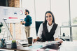 © deagreez - Businesswoman smiling at desk with charts in modern loft office while colleague presents in background