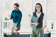 © deagreez - Confident businesswoman in bright office smiles at camera holding folder with colleague working nearby