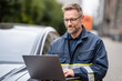© Kowit - A man in a work uniform uses a laptop while standing next to a car outdoors, focused on his task.