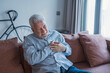 © Daniel - An older man experiences chest pain while sitting on his couch at home, showing signs of a heart attack and medical distress in a quiet and vulnerable moment