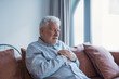 © Daniel - An elderly man clutches his chest while seated on the couch, suffering severe pain that suggests a cardiac emergency, highlighting the danger of heart disease at home