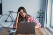 © Daniel - An overworked woman sits at her workspace beside a laptop, pressing her hands to her head as she reacts to negative news from work or bank emails, showing anxiety and emotional exhaustion
