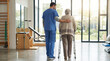 © Cliff - Rear view of male physiotherapist supporting senior woman with walker frame during rehabilitation session in bright modern clinic, geriatric care and recovery concept