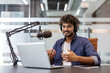 © Tetiana - Smiling Indian man wearing headphones sitting at a desk in the office in front of a microphone and talking on a video call on a laptop