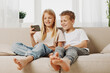 © SHOTPRIME STUDIO - Two happy children, boy and girl, sitting barefoot on a couch, smiling while looking at a smartphone together in a bright living room. Concept of childhood, technology, and friendship.