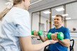 © wedmoments.stock - Senior man performing supervised strength training exercises with green dumbbells while sitting in a rehabilitation center with a female trainer assisting him