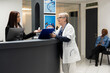 © DC Studio - Asian administrator discusses reports and scheduled appointments with elderly general practitioner at front desk. Senior doctor holds clipboard, reviewing patient registrations with receptionist.