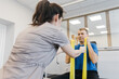 © wedmoments.stock - Patient performing upper body rehabilitation exercises with resistance bands while being assisted by a healthcare professional in a bright therapy room