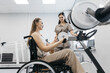 © wedmoments.stock - Female patient in wheelchair using rehabilitation exercise equipment with a smiling therapist assisting in a bright, modern therapy room