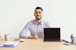 © Prostock-studio - A man sits at a wooden desk with a laptop and smartphone. He smiles directly at the camera. The office has bright lighting and minimal decoration, creating a simple workspace.