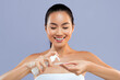 © Prostock-studio - A woman stands with a towel around her upper body, smiling as she pumps skincare product into her hand. The background is purple, creating a cheerful atmosphere as she prepares to apply the product.