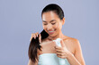 © Prostock-studio - A woman with long hair is applying a hair product to her hair while smiling. She is wearing a towel and stands in a studio with a plain gray background. Her expression shows happiness and confidence.