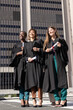 © wavebreak3 - Diverse female graduates posing on plaza by high-rise, wearing black gowns, holding rolled diplomas
