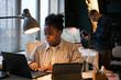 © DragonImages - Black woman working on laptop at desk in modern office, focusing on screen while typing, with Caucasian man standing in background holding camera and adjusting equipment