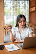 © bongkarn - Pretty asian woman holding pen talking to video call on laptop at wooden table while sitting in cafe
