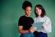 © we.bond.creations - Two smiling women from diverse backgrounds collaborating and sharing ideas while using a portable digital tablet device against a green background