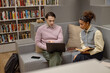 © Seventyfour - Young adult Caucasian man explaining information on laptop to young adult woman holding book, both sitting on sofa in library surrounded by bookshelves and study materials