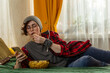 © _KUBE_ - Caucasian teenage woman student resting indoors during free time. Girl eats popcorn and using smartphone. Home comfort theme. Mid shot