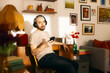 © Francis - A relaxed man with a shaved head and tattoos sits in a sunlit, vintage-style living room, wearing over-ear headphones and looking at his smartphone while enjoying music or a podcast.