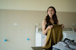 © Tj - Young Asian woman smiling, holding up a clean green sweater, and organizing a pile of freshly washed laundry items on a countertop in a bright home utility room