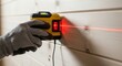 © MAJIDULISLAM - closeup of a hand holding a yellow laser level projecting a red beam onto a white wooden wall for precise measurement and alignment