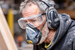 © Kowit - A close-up of a bearded man wearing protective goggles, a respirator mask, and earmuffs while working in a workshop.