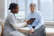 © Seventyfour - Black young adult woman nurse showing digital tablet to Caucasian middle aged woman patient during medical consultation in clinic examination room, both focused on screen