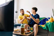 © kleberpicui - Brazilian father and teenage son watching football match together at home celebrating exciting moments. Family bonding through sports entertainment sharing emotion, support and generational connection