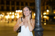© JackF - Young smiling woman at Royal square, leaning on pole, spend time in public urban space. Electric street lighting, twilight in southern city, evening life in big city