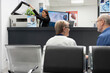 © DC Studio - Asian receptionist using copier and scanning machine at front desk of modern healthcare facility. Female hospital secretary handles printing of medical forms as elderly doctor consults with patient.