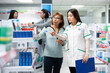 © DC Studio - Medicine consultant pointing at pharmaceutical products on shelves, giving detailed information to curious client in modern drugstore. Asian women looking at medication packages in pharmacy aisle.