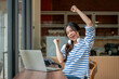 © bongkarn - Excited asian woman looking at laptop raising both fist as sitting at wooden counter table in a cafe