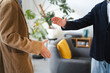 © Studio Marmellata - Two men in casual jackets shake hands, signifying a greeting or agreement in a modern office setting with plants and furniture.