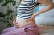 © wifesun - Pregnant woman sits on a bed, holding her lower back with one hand and her baby bump with the other, clearly experiencing discomfort and pain associated with pregnancy while anticipating motherhood